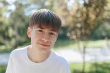 Basit beyaz gömlek giymiş neşeli bir ergen çocuk, açık havayı seviyor. Güneşli park çevresi portre için taze ve doğal bir zemin sağlıyor..