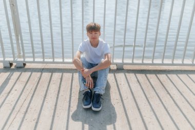 Nehrin kenarında metal bir korkulukta oturan, kot pantolon ve beyaz tişörtlü genç bir çocuk, güneşli bir günde sakin ve rahat bir anın tadını çıkarıyor.