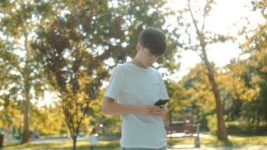 Güneşli bir günde şehir parkında beyaz tişörtlü, akıllı telefonlu genç bir çocuk. Etrafı yeşilliklerle çevrili, gençlik neşesi ve özgürlük yayıyor.