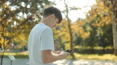 Güneşli bir günde şehir parkında beyaz tişörtlü, akıllı telefonlu genç bir çocuk. Etrafı yeşilliklerle çevrili, gençlik neşesi ve özgürlük yayıyor.