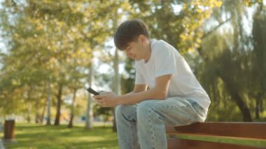 Beyaz tişörtlü, elinde akıllı bir telefon olan genç bir çocuk güneşli bir günde şehir parkında parlak bir şekilde gülümsüyor. Etrafı yeşilliklerle çevrili, gençlik neşesi ve özgürlük yayıyor.