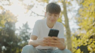 Beyaz tişörtlü, elinde akıllı bir telefon olan genç bir çocuk güneşli bir günde şehir parkında parlak bir şekilde gülümsüyor. Etrafı yeşilliklerle çevrili, gençlik neşesi ve özgürlük yayıyor.