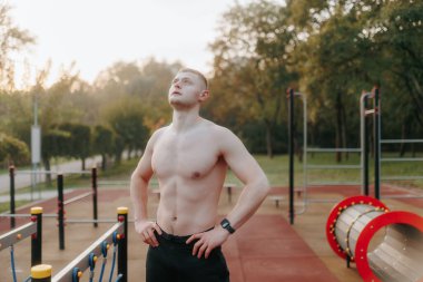 Tişörtsüz genç adam dışarıda çalışırken biraz mola verir. Kaslı vücudu ve kararlı ifadesi aktif yaşam tarzını ve uygunluğa olan bağlılığını vurguluyor..