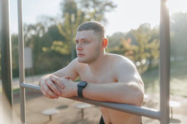 Genç, kaslı, çıplak gövdeli bir adam açık hava spor salonunda dinleniyor. Yoğun bir antrenmandan sonra yorgun görünüyor. Sağlıklı, aktif bir yaşam tarzı var..