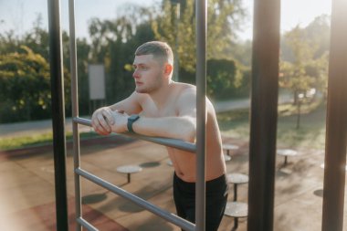 Çıplak gövdeli formda bir genç, spor salonunda biraz dinlenir. Kaslı vücudu ve yorgun ifadesi vücut çalışmasının yoğunluğunu gösterir..