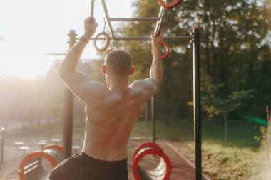 Genç, zinde bir adam arkadan görülüyor, çıplak gövdesi jimnastik yaparken parıl parıl parıl parlıyor açık bir spor salonunda yüzüklerle. Sahne gücü yakalar ve odaklanır..