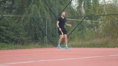 Siyah spor giyimli bir adam kırmızı tenis kortunda topa vurmaya hazırlanıyor. Çevreleyen ağaçlar ve bulutlu gökyüzü bu açık hava sportif anını çerçeveliyor..