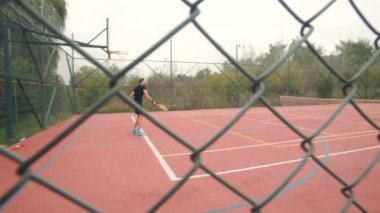 Siyah tişörtlü ve şortlu bir oyuncu kırmızı tenis kortunda topa vurmaya hazır. Açık hava maçı yeşilliğin ortasında ve bulutlu bir gökyüzünün altında yapılıyor..