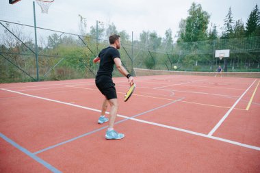 Siyah spor giyimli bir adam kırmızı tenis kortunda topa vurmaya hazırlanıyor. Çevreleyen ağaçlar ve bulutlu gökyüzü bu açık hava sportif anını çerçeveliyor..