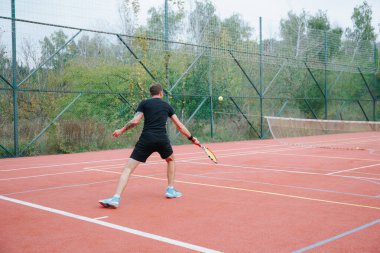 Eylemin ortasında yakalanan siyah elbiseli bir erkek tenisçi raketini kırmızı tenis kortunda sallıyor. Çevreleyen ağaçlar ve bulutlu gökyüzü doğal bir zemin hazırladı.