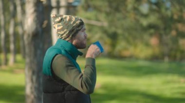 Açık hava kıyafetleri içinde kamuflaj beresi de dahil olmak üzere bir adam, huzurlu bir orman sahnesinin ortasında durup doğal çevreyi kucaklarken mavi bardaktan içmekten zevk alıyor..