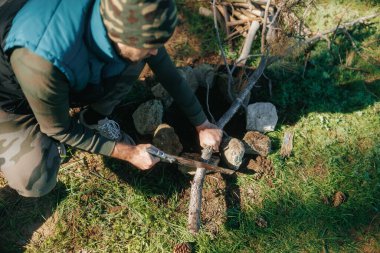 Huzurlu bir ormanda odun kesmek için el testeresi kullanan bir adam kamp macerasına hazırlanıyor. Doğayla iç içe bir kamp ateşi için dallar topluyor..