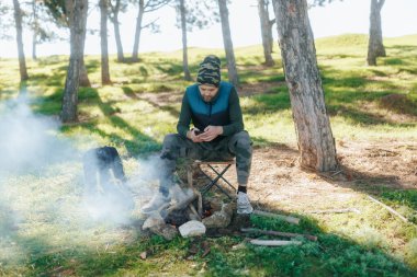 Vahşi doğada kamp ateşinin yanında akıllı telefon kullanan bir adam oturuyor. Doğa ile çevrili, açık hava macerasıyla modern teknolojiyi barışçıl bir ortamda birleştiriyor..
