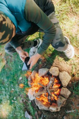 A camper sits by the warmth of a campfire, using his smartphone. The image combines rugged wilderness survival with the ease of modern digital life. clipart