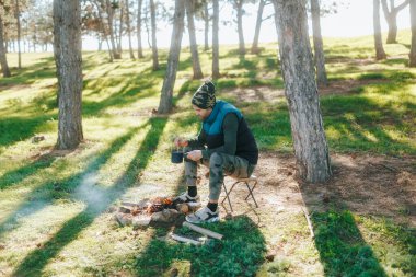 Kamuflaj kıyafetli bir adam kamp ateşinin üzerinde bir demlik karıştırıyor, ormanın sıcak ve sakin ortamının tadını çıkarıyor. Rahatlamak için mükemmel bir yer..