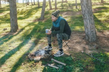 Barışçıl bir açık hava ortamında bir adam elinde sıcak bir bardakla küçük bir kamp ateşinin yanında oturur. Macera için giyinmiş, sakin bir sabahta doğanın huzurunu kucaklıyor..