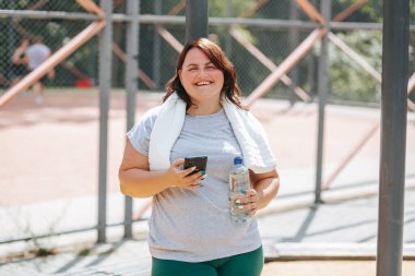 Mutlulukla dolu obez bir kadın, eğitimden sonra akıllı telefonunu kontrol ediyor. Yüzünde bir memnuniyet gülümsemesi, elinde bir su şişesi tutuyor..