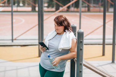 Açık hava egzersizinden sonra, kilosu büyük bir dişi akıllı telefonunu kullanıyor. Mutluluğu, bir su şişesi ile tatmin gülümsemesiyle aşikar..