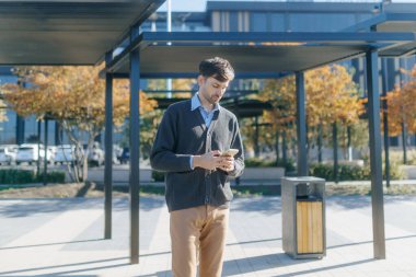 Sıradan giyinmiş bir adam şehir istasyonunda tek başına duruyor, saati takip etmek için akıllı telefonunu kontrol ediyor, sakin ve düşünceli bir bekleyiş anını somutlaştırıyor..