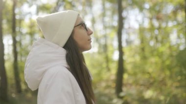 Soğuk hava için giyinmiş, gözlüklü bir kadın ormanda duruyor, yumuşak sonbahar ışığı altında, ağaçlarla çevrili, ve doğayı kucaklayan sakin bir atmosfer..