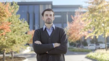 İş kıyafetleri giymiş bir adam kolları bağlı, açık bir sonbahar gününün tadını çıkarıyor. Yakındaki ağaçların ılık renkleri mevsimlik bir dokunuş ekliyor.