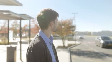 Sıradan giyinmiş, düşünceli bir adam, sonbahar renkleri ve modern mimariyle çevrili, sakin bir bekleyiş anını somutlaştıran bir şehir istasyonunda duruyor..