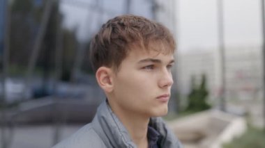 Yüzünde ciddi bir ifade olan genç bir çocuğun, şehir ortamında dikilirken, modern binaların arka planına karşı sıradan bir ceket giymesi..