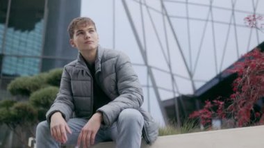 Şehir ortamında oturan genç bir adam, gri bir ceket ve kot pantolon giyiyor, şehirde huzurlu ve kaygısız bir ifadeyle boş zamanlarının tadını çıkarıyor..