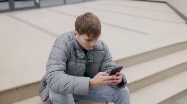 Genç adam dışarıda oturuyor, kulaklık takmış ve elinde telefonla boş zamanın tadını çıkarıyor. Huzurlu bir şehir anı yakalıyorum. Dijital dinlenme ve rahatlama..