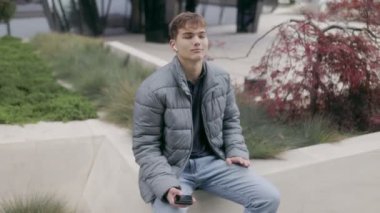 Sakin bir ifadeyle, kulaklıkla müzik dinlemekten ve şehir ortamında telefon tutmaktan hoşlanan genç bir adam. Modern bir şehir sahnesinde sakin ve tasasız.