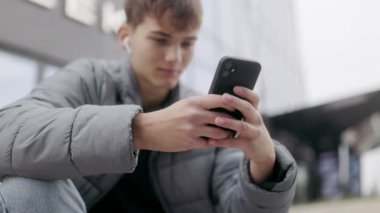 Şehirdeki modern gençlik sahnesinde bir genç oturuyor ve elinde telefonla müzik dinliyor. Sakin, rahat ve açık şehir ortamında rahat..