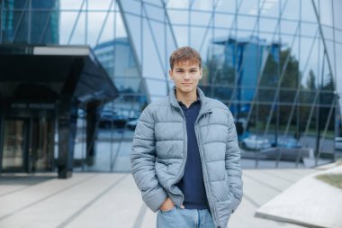 Kabarık bir ceket giyen ergen, kentte kendine güvenen ve modaya uygun, rahat bir tarz sergileyen modern bir şehir zeminine poz veriyor..