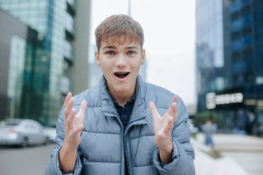 Young man with wide eyes and open mouth expresses surprise in an urban environment. His raised hands and city background add emphasis to his genuine reaction. clipart