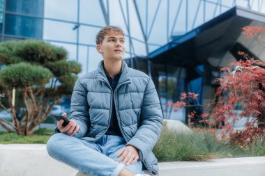 Youthful, carefree moment of a teenager enjoying music on headphones and relaxing with his smartphone in a city setting, capturing modern digital leisure. clipart