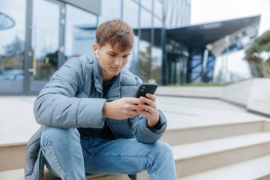 Young male enjoying leisure time, sitting with his smartphone and listening to music. Casual moment of youth in an urban background with modern architecture. clipart