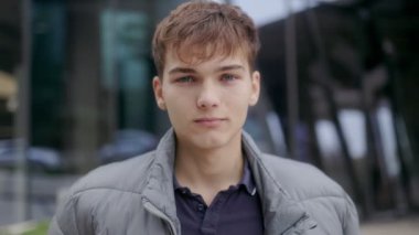 Teçhizatlı gülümseyen bir gencin portresi, arka planda modern bir binayla ayakta duruyor, kendine güveni ve gençlik cazibesi yayıyor..