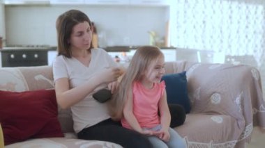 Genç bir kız, annesinin uzun saçlarını tararken sıcak bir gülümsemeyle evlerindeki rahat bir kanepede otururken aile sevgisinin hassas bir anını yakalıyor..