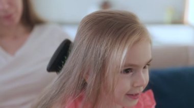 Bir anne, sıcak bir evde otururken kızının saçlarını nazikçe tarar. Aile içinde sevgi ve ilgi dolu bir an yaşar..