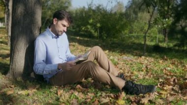 Bir adam çimlere oturur, ağaca yaslanır ve kitap okur. Sonbahar yaprakları ve huzurlu açık hava sakin ve yansıtıcı bir anı yakalıyor..