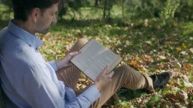 Kitap okurken el ele tutuşan, ağacın yanındaki çimlerin üzerinde oturan bir adamın yakın çekimi. Huzurlu ortam, dökülen yapraklar ve sonbahar renkleri sakin ve yansıtıcı bir atmosfer yaratır..
