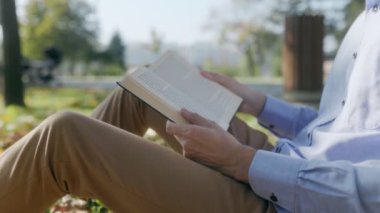Sonbahar yaprakları ve çimenli çevresi manzaralı bir kitap tutan bir adamın yakın çekimi. Bir ağacın yanında otururken, doğada bir anlık yalnızlık ve rahatlığın tadını çıkarıyor..