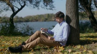 Düşünceli orta yaşlı bir adam gölün kenarındaki bir ağacın altında oturup kitap okuyor ve sonbahar yapraklarıyla çevrili güneşli bir sonbahar gününde doğanın huzurunun tadını çıkarıyor..