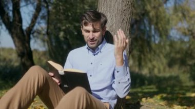 Sakallı ve yüzünde şaşırmış bir ifade olan meraklı bir öğrenci huzurlu bir sonbahar parkında kitap okuyor. Sıcak güneş ışığı ve renkli yapraklar dingin bir zemin oluşturur..