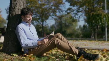 Bir adam bir ağaca yaslanır, bir kitapta kaybolur, sonbahar güneşi ışıl ışıl parlarken. Huzurlu park yeri rahatlama ve neşe dolu mükemmel bir an sağlıyor..