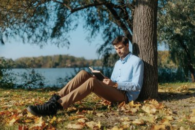 Düşen yaprakların arasında, sıradan giyinmiş bir adam ağaca yaslanırken kitap okur. Sahne, açık havada sessiz ve iç gözlemci bir sonbahar anını yansıtıyor..