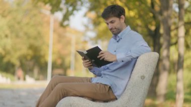 Günlük kıyafetli bir adam parkta bir bankta oturur, kameraya gülümser ve bir kitap gösterir, ağaçlar ve güneş ışığının içinden süzüldüğü sakin bir sonbahar sahnesine kuruludur.