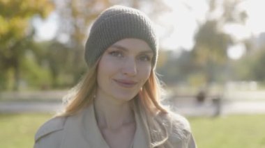 Sonbahar için giyinmiş bir kadın, yumuşak bir gülümsemeyle, renkli yapraklarla ağaçların arasında duruyor, güneşli bir günün tadını çıkarıyor huzurlu bir ortamda..