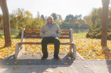 Senior man with beard and glasses sits peacefully on a park bench, surrounded by autumn colors. The serene setting offers a moment of relaxation and reflection. clipart