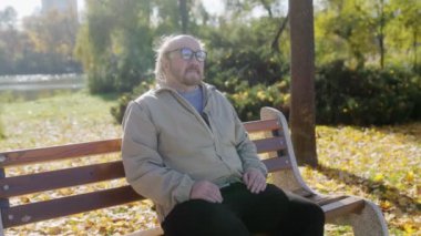 Sakallı ve gözlüklü yaşlı adam sonbahar renkleriyle çevrili bir bankta huzur içinde oturuyor. Huzurlu ortam bir rahatlama ve yansıma anı sunar..