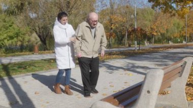 Gözlüklü yaşlı bir adam dışarıda kalp sorunu yaşıyor, ve hemşiresi ona yardım ediyor, bir bankta oturmasına yardım ediyor..